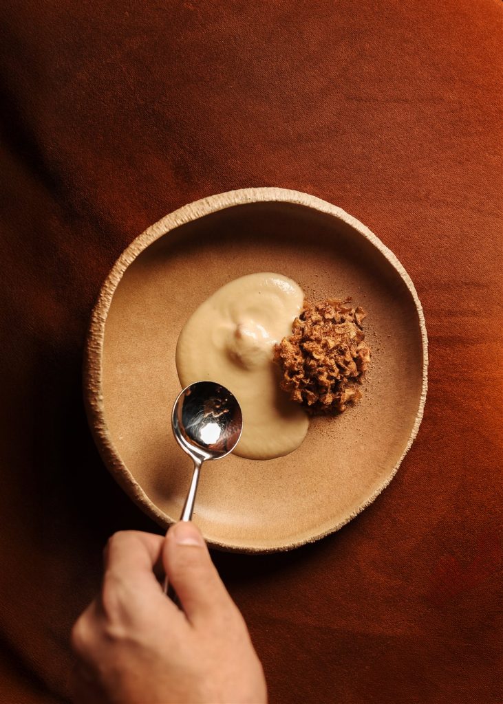 Chef's hand and spoon over a wooden bowl with sauce and grain — intimate cooking moment from Mattia Stanchieri's work