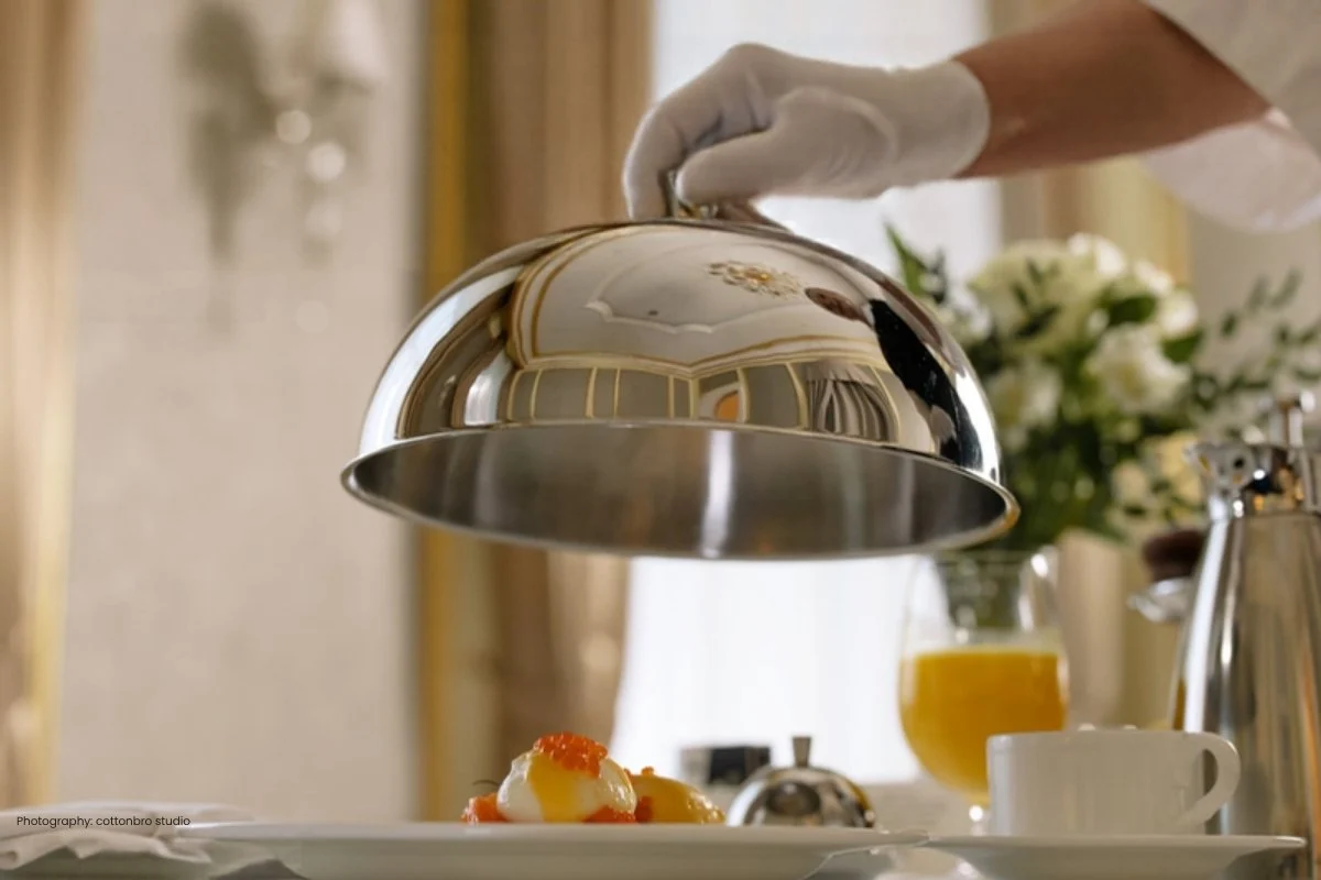 A white-gloved waiter lifts a silver cloche at a luxury hotel table